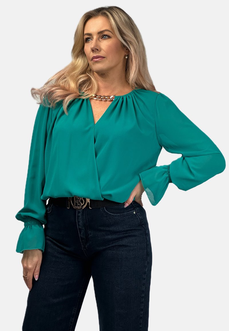 Turkusowa jedwabna bluzka z drapowanym dekoltem w serek, bufkami i marszczonymi detalami na ramionach. Połączona z ciemnymi dżinsami i paskiem z złotymi elementami.