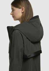 Giacca impermeabile verde scuro con cappuccio, chiusura con zip e una striscia con logo in rilievo sulla schiena. Presenta una superficie liscia e un design aderente.