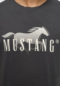 Camiseta de algodón negra con un gráfico de un caballo galopante en blanco y el texto "MUSTANG" debajo en letras mayúsculas y negritas. Textura suave.