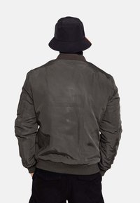 Olivgrüne Bomberjacke mit geripptem Kragen und Bündchen, die eine glatte Textur und einen entspannten Schnitt aufweist. Getragen mit einem schwarzen Bucket Hat.