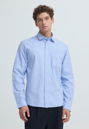 Ung mand iført en lyseblå skjorte med vertikale striber og knapper, lange ærmer, stående foran en ensartet lys baggrund.