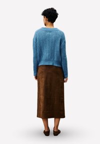 Une personne avec des cheveux courts et bouclés porte un pull en maille bleu, une jupe midi marron et des chaussures à enfiler marron, se tenant de dos.