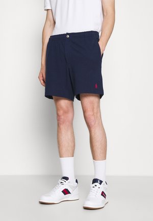 Uomo che indossa pantaloncini blu navy con elastico in vita e sneaker bianche con dettagli blu navy e rossi, in piedi davanti a uno sfondo semplice.