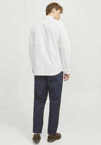 Camisa blanca de manga larga con cuello abotonado, combinada con pantalones azul oscuro y zapatos marrones. La camisa cuenta con un dobladillo curvado.