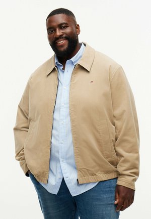 Smilende mand iført en beige jakke over en lyseblå skjorte med knapper og blå jeans, stående med hænderne i lommerne mod en hvid baggrund.