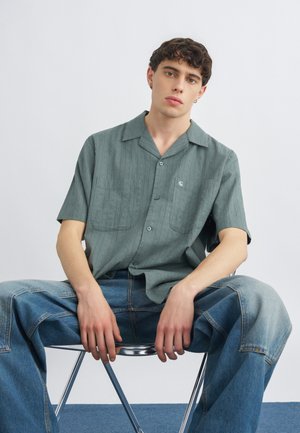 Νεαρός άνδρας με σγουρά μαλλιά κάθεται σε μεταλλική καρέκλα φορώντας πράσινο πουκάμισο με κοντά μανίκια και ανοιχτά μπλε τζιν, πάνω σε απλό φόντο.