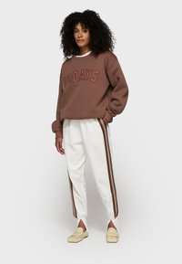 Brauner Baumwollpullover mit "10 DAYS" in dunklerem Braun bedruckt. Cremefarbene Hose mit braunen Seitenstreifen und Schlitzen an den Bündchen. Beigefarbene Slipper.