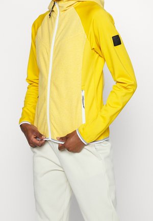 Personne portant une veste jaune zippée avec une fermeture éclair blanche et des accents de poche, et un pantalon clair, ajustant la fermeture éclair de la veste.