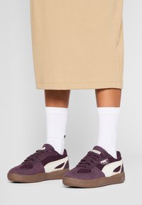 Puma-sneakers med ett djupt lila mockaskinn, vita sidodetaljer, gummisula och texturerad nät-tunga, tillsammans med vita strumpor.