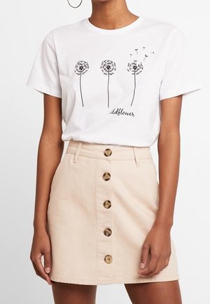 Kvinde iført en hvid t-shirt med tre sorte mælkebøtter og beige nederdel med knapper foran.