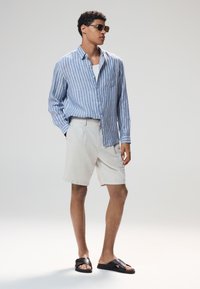 Chemise légère à rayures bleues et blanches avec boutons, short beige et sandales noires. Le modèle porte des lunettes de soleil et a les cheveux courts et bouclés.