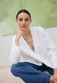 Femme aux cheveux bruns tirés en arrière portant un blazer blanc texturé et un jean bleu, reposant le menton sur la main devant un fond vert et beige.