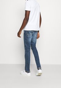 Istuvat siniset farkut, joissa lievä haalistuma, taustataskuissa ompeluaksentteja, yhdistettynä valkoiseen lyhythihaiseen paitaan ja valkoisiin lenkkareihin.