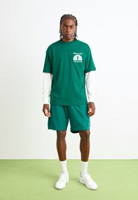 Grøn bomulds t-shirt med hvidt logo, parret med matchende grønne shorts. Modellen har en hvid langærmet bluse og hvide sneakers på. Ensartet baggrund.