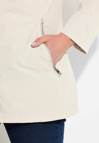 Světle béžová bunda s hladkým povrchem, vybavená boční kapsou na zip a rovně střiženým lemem, doplněná o tmavě modré džíny.