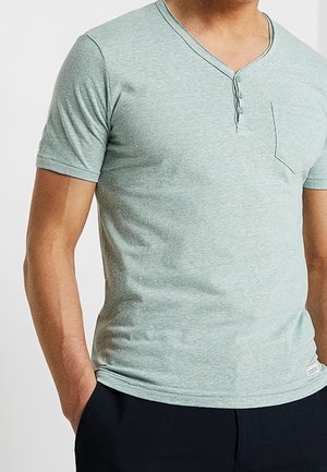 Mężczyzna w jasnozielonej koszulce typu henley z krótkim rękawem i kieszenią oraz czarnych spodniach, stojący z jedną ręką w kieszeni.