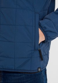 Navy quilted jacket with a smooth texture, featuring a zippered pocket, elastic cuffs, and a logo tag at the hem.