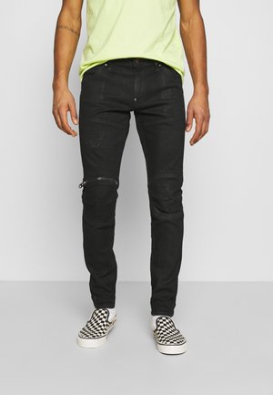 Homme portant un jean noir slim avec des fermetures éclair aux genoux et des détails usés, une chemise jaune néon et des chaussures slip-on à carreaux noir et blanc.