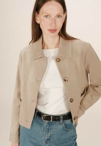 Donna con capelli castani lisci indossa una giacca beige con bottoni sopra una maglietta bianca e jeans blu con cintura nera su uno sfondo neutro.