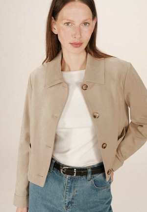 Donna con capelli castani lisci indossa una giacca beige con bottoni sopra una maglietta bianca e jeans blu con cintura nera su uno sfondo neutro.