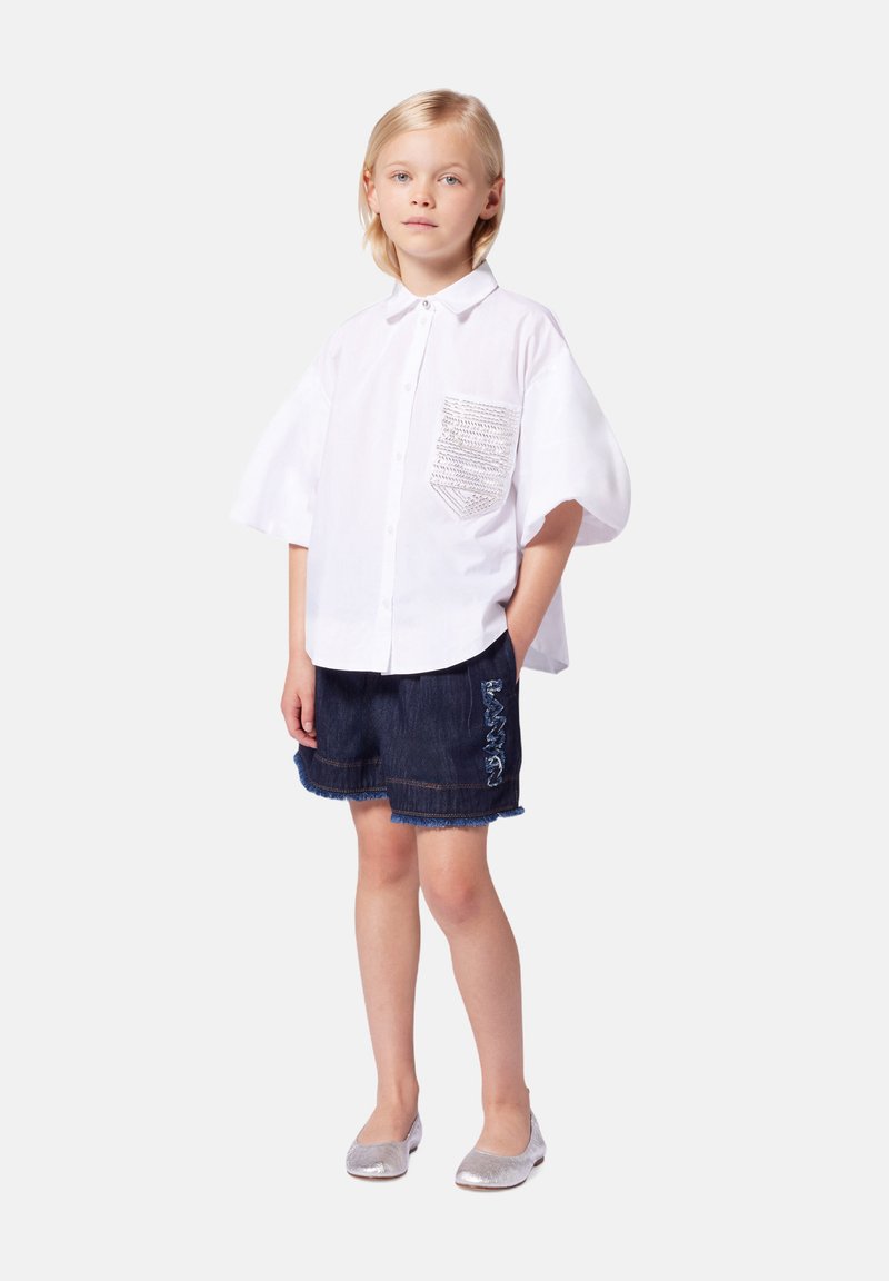 Chemise blanche à manches courtes avec poche texturée, associée à un short en denim sombre avec ourlet effiloché et détail brodé bleu. Ballerines argentées.