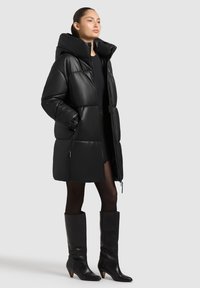 Žena oblečená v čiernej oversized prešívanej bunde, čiernej sukni, priesvitných pančuchách a čiernych kožených čižmách po kolená na svetlošedom pozadí.