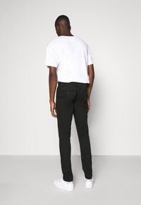 Pantalons noirs en tissu lisse, Coupe slim, dotés de poches arrière et de coutures contrastées, associés à un T-shirt blanc uni.