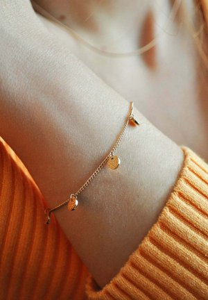 Goldene Kettenarmband mit strukturierten kreisförmigen Anhängern, getragen an einem Handgelenk gegen einen orangefarbenen gerippten Pullover. Glatte und glänzende Oberflächendetails.