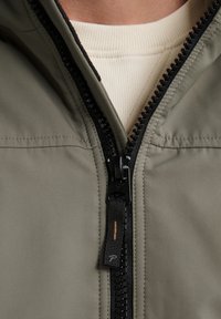 Giacca khaki con zip, caratterizzata da una superficie testurizzata, zip nera e dettagli di cucitura visibili. Lo strato interno è in un tessuto di colore crema.