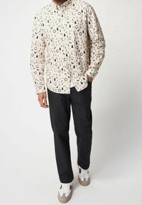 Camicia a maniche lunghe con base crema, decorata con un motivo di ghirlande verdi, alberi e bastoncini di zucchero, abbinata a pantaloni neri e sneakers.