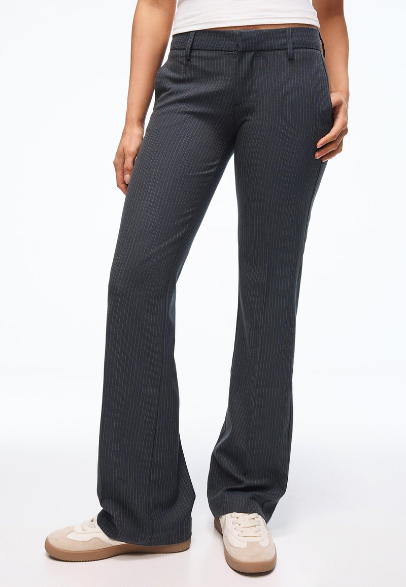 Femme portant un pantalon large à rayures fines gris foncé et des baskets blanches, debout devant un fond blanc uni.
