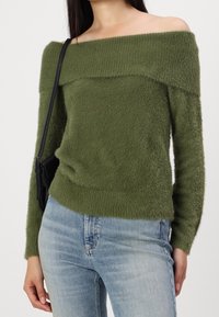 Maglione verde scollato sulla spalla con una superficie texturizzata e soffice. Maniche lunghe e orlo rotondo. Indossato con jeans in denim azzurri.