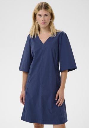 Kobieta o blond włosach, ubrana w niebieską sukienkę do kolan z szerokimi rękawami do łokcia i perłowymi kolczykami, stojąca na jednolitym tle.