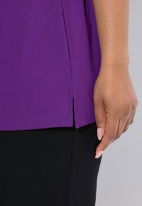 Paarse shirt met een zijsplit aan de zoom, gemaakt van glad materiaal. Hand rustend tegen een zwarte stof, waarbij de natuurlijke huidtextuur zichtbaar is.