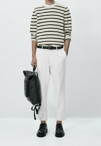 Gestreiftes Langarmhemd in Creme und Schwarz, weiße Hose, schwarze Lederschuhe und ein schwarzer Rucksack mit Reißverschlüssen und verstellbaren Trägern.