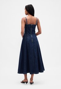 Tamnoplava traper maksi haljina s podesivim naramenicama, pripijenim korzetom, širokom suknjom i središnjim patentnim zatvaračem na leđima. Glatka tekstura s vidljivim šavovima.