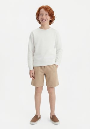 Χαμογελαστός κοκκινομάλλης αγόρι που φοράει λευκό πουκάμισο με μακριά μανίκια, μπεζ σορτς και καφέ παπούτσια τύπου slip-on, στέκεται με το ένα χέρι στην τσέπη.