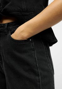 Käsi on sisestatud mustade denim-pükste esi-taskusse, näidates õmmeldud õmblusi ja osa sobivast mustast jakkist valge taustaga.