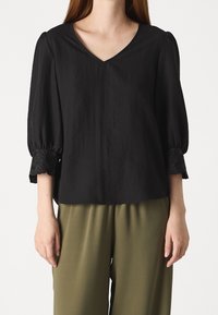 Blusa nera con scollatura a V, maniche a sbuffo e pizzo sui polsini; abbinata a pantaloni larghi color verde oliva. Tessuto leggero.