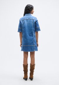 Φόρεμα πουκάμισο από τζιν σε μεσαίο μπλε, με κοντά μανίκια, κανονικό γιακά και ραφές σε αντίθεση. Συνδυάζεται με καφέ σουέτ μπότες μέχρι το γόνατο.