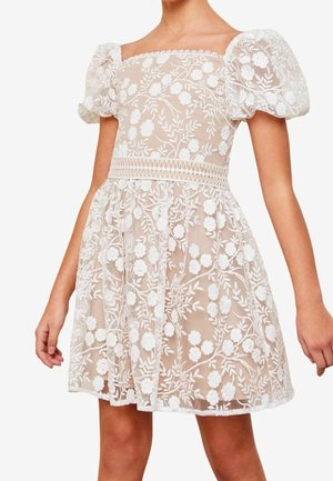 Krótka biała sukienka z kwiatowej koronki z bufkami i kwadratowym dekoltem, noszona przez osobę o opalonej skórze bez widocznych dodatków.