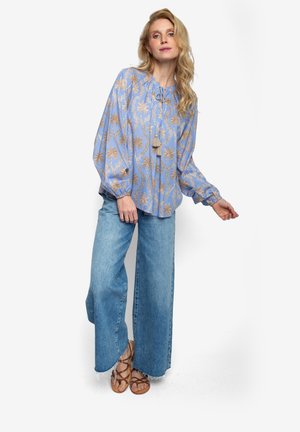 Jasnoniebieska bluzka w złoty liściasty wzór, luźny krój, wiązanie przy dekolcie i długie rękawy. W połączeniu z szerokimi dżinsami o szerokich nogawkach i brązowymi sandałami.