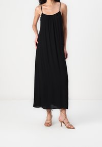 Černé maxi šaty s tenkými ramínky, volným střihem a nařasenou vrchní částí. Modelka má na sobě béžové sandály na tenkých podpatcích, což ukazuje na ležérní, volný siluetu.