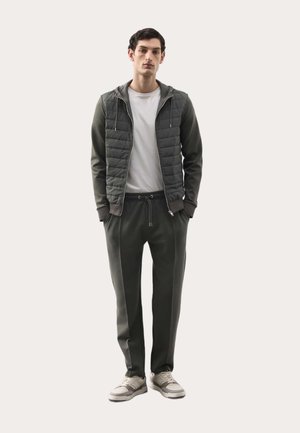 Mand iført en mørkegrøn quiltet lynlåstrøje, hvid t-shirt, matchende mørkegrønne bukser og beige sneakers, stående med hænderne i lommerne.