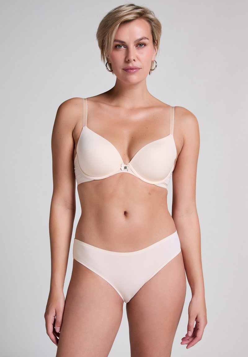 Vrouw met kort blond haar, gekleed in een lichtbeige bh en bijpassend ondergoed, staand tegen een effen lichtgrijze achtergrond.