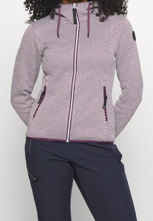 Mujer con chaqueta con capucha y cremallera con un patrón geométrico blanco y morado y pantalones azul marino oscuro con bolsillos con cremallera.