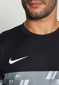 Muž v černém a šedém sportovním tričku značky Nike s bílým logem Swoosh, zobrazující spodní část obličeje a krk s upraveným vousem.