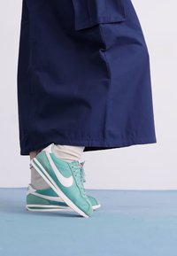 Nike sneakers in mint green with white swoosh, paired with light-colored socks, beneath a long navy blue garment. Smooth surface and classic design.