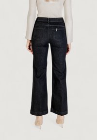 Nainen, jolla on tummat leveät housut ja beigeet korkokengät, näkyy selkäpuolelta yksinkertaista taustaa vasten.