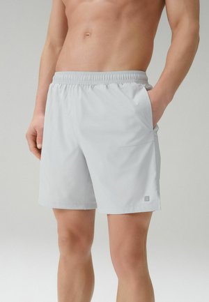 Férfi világosszürke, elasztikus derekú rövidnadrágban, kezét a zsebében tartva, meztelen törzzsel és lábbal egyszínű háttér előtt.